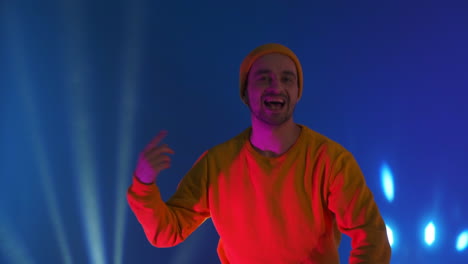 Ein-Mann-Mit-Gelbem-Kapuzenpullover-Und-Hut-Tanzt-Auf-Blauem-Hintergrund.-Lustiges-Tanzen-Beim-Blick-In-Die-Kamera